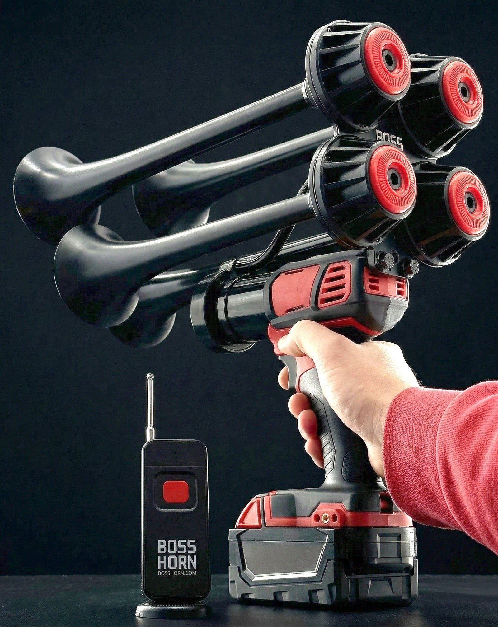 Person aiming the black-and-red four-trumpet train horn upward by the handle, remote and battery in foreground on a dark background - view 12