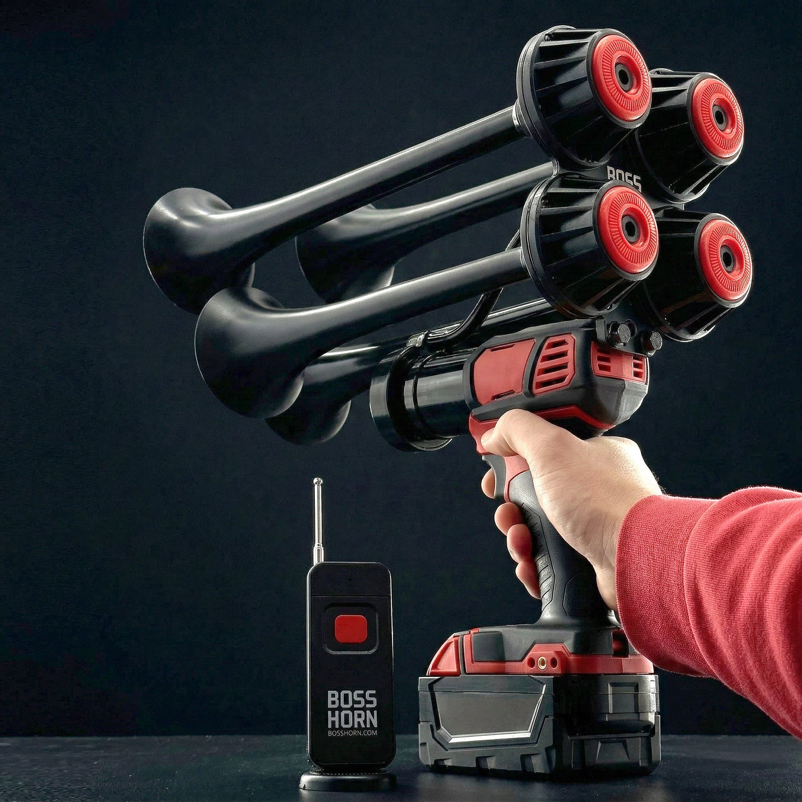 Hand gripping the train horn by its trigger handle, aiming the black-and-red quad trumpets upward, with remote and battery on a dark surface - view 9