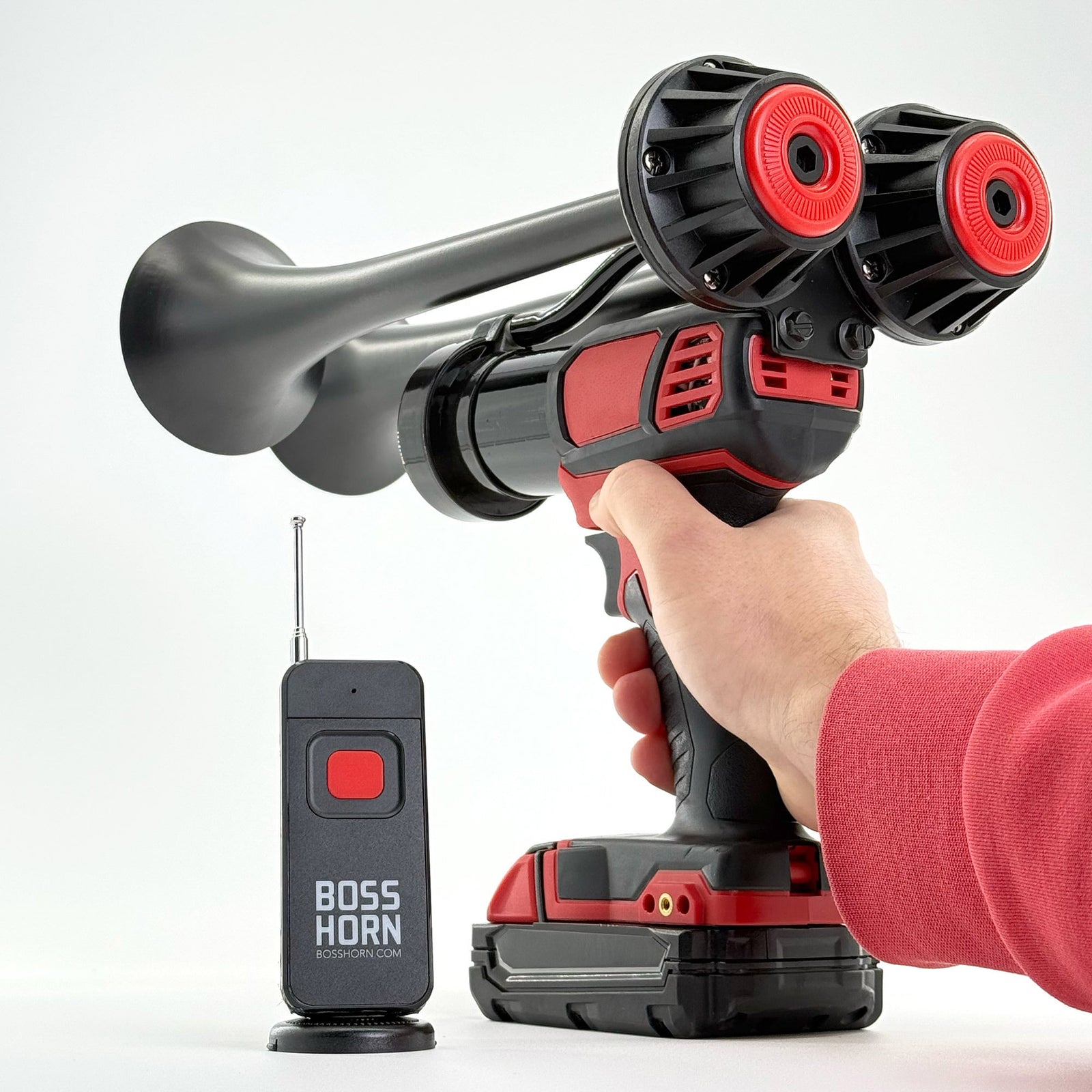 Hand holding and aiming the black dual-trumpet train horn on a red cordless power tool with Milwaukee® M18 battery, remote nearby on white - view 9